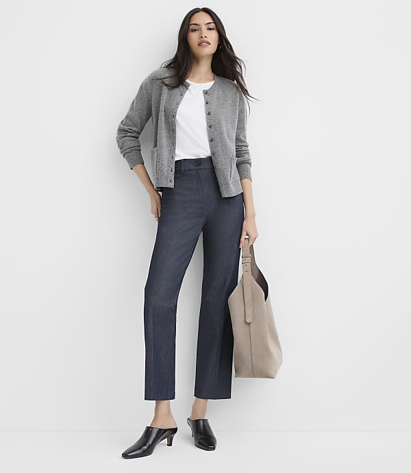 A woman wearing a grey cardigan, white t-shirt, and navy trousers stands holding a beige tote bag against a plain white background.