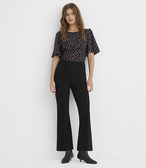 A woman in a floral top and black flared pants stands against a plain white background.