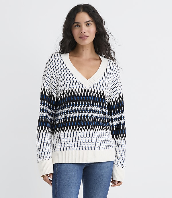 A woman wearing a patterned white and blue V-neck sweater and blue jeans stands against a plain white background.