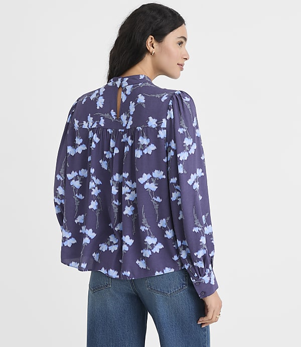A woman wearing a purple floral blouse and blue jeans stands against a plain white background.