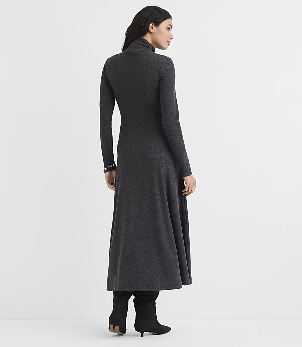 A woman wearing a long-sleeved, charcoal grey maxi dress and black boots stands against a plain white background.