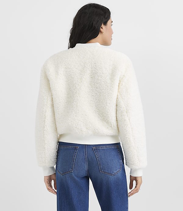 A woman wearing a white fuzzy jacket and blue jeans stands with her back to the camera against a plain light gray background.