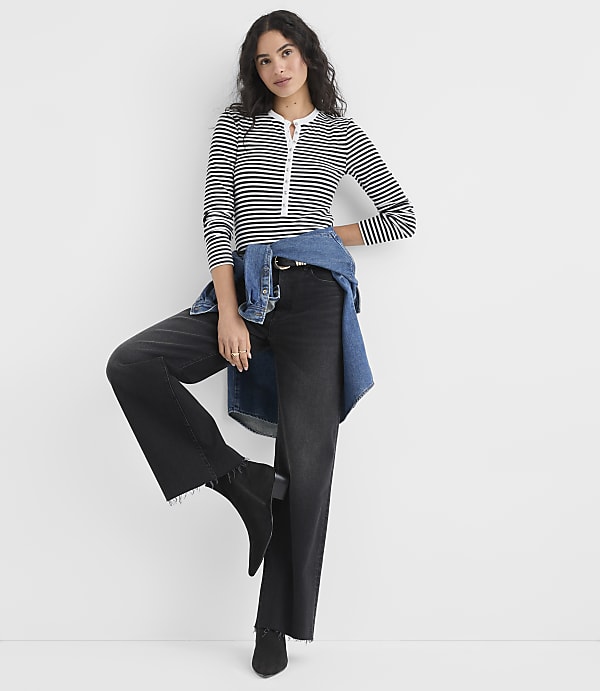 A woman wearing a striped shirt and wide-leg black pants with a denim shirt tied around her waist stands against a plain white background.
