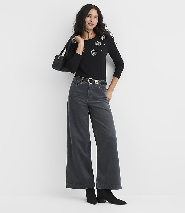 A woman wearing a black sequined top, wide-leg grey trousers, and black boots stands against a plain white background.