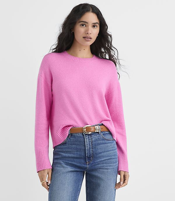 A woman wearing a bright pink sweater and blue jeans stands against a plain white background.
