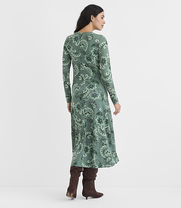 A woman wearing a green paisley print maxi dress and brown boots stands against a plain white background.