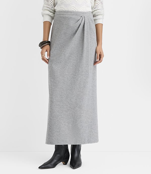 A person wearing a white sweater, a long grey skirt, and black boots stands against a plain white background.