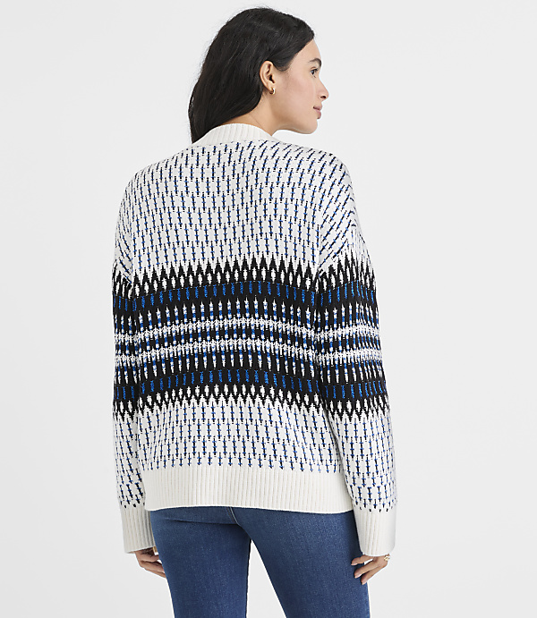 A woman wearing a patterned white, blue, and black sweater and blue jeans stands against a plain white background.