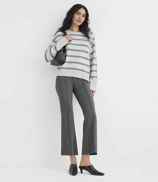 A woman wearing a grey striped sweater, dark grey trousers, and black heels stands against a plain white background.