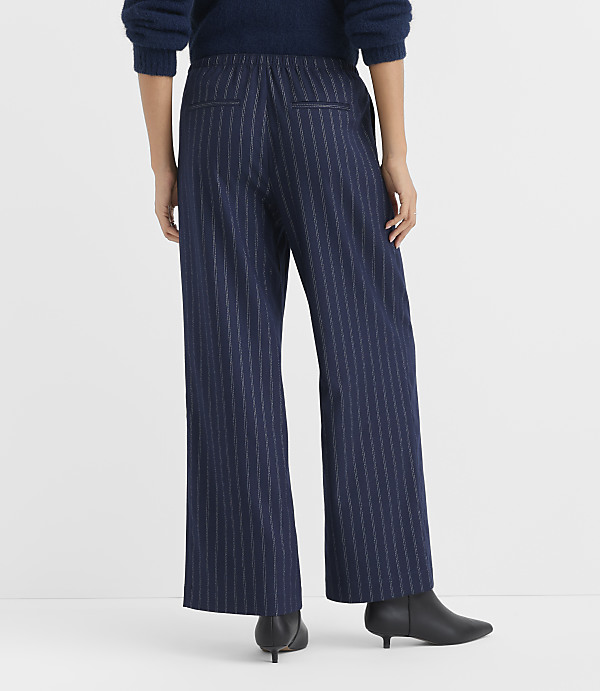 A person stands facing away from the camera wearing a blue sweater, navy pinstriped wide-leg trousers, and black boots against a plain white background.