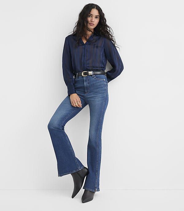 A woman wearing a navy blue striped blouse, flared blue jeans, and black pointed boots stands against a plain white background.