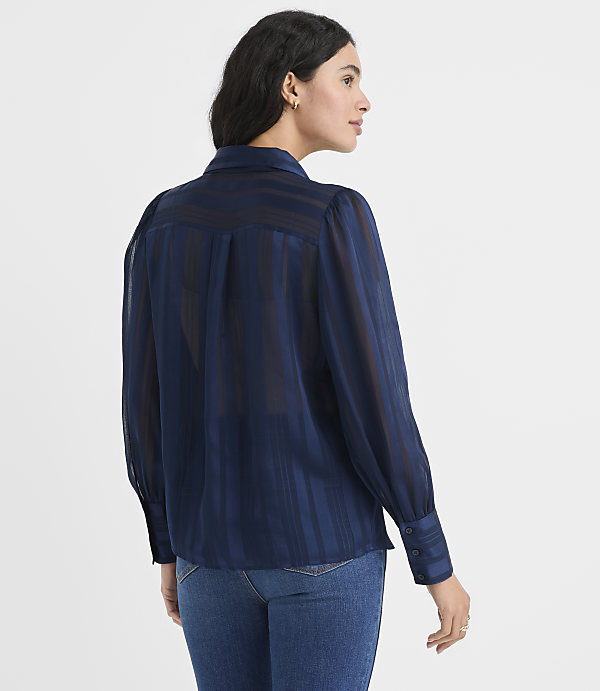 A woman wearing a navy blue striped blouse and blue jeans stands against a plain white background.