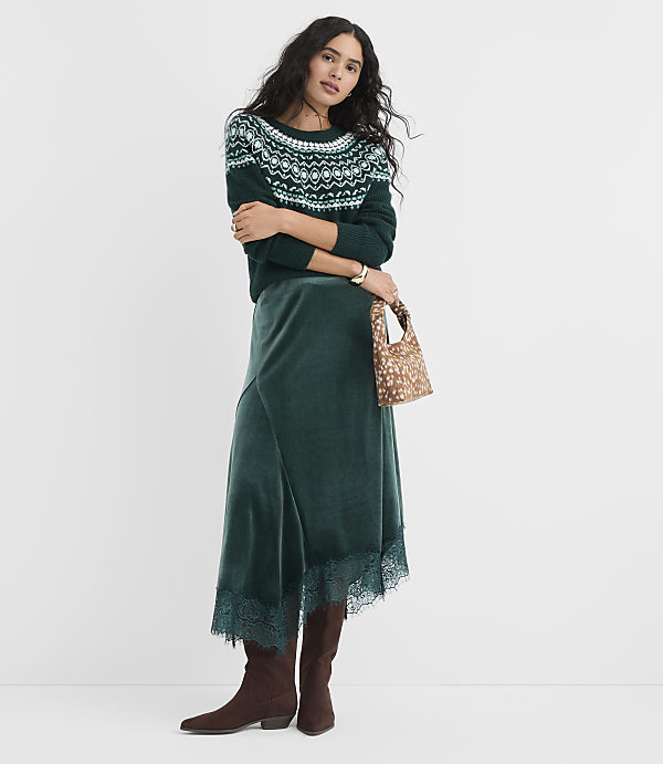 A woman wearing a green patterned sweater, a matching green velvet skirt, and brown boots stands against a plain white background.