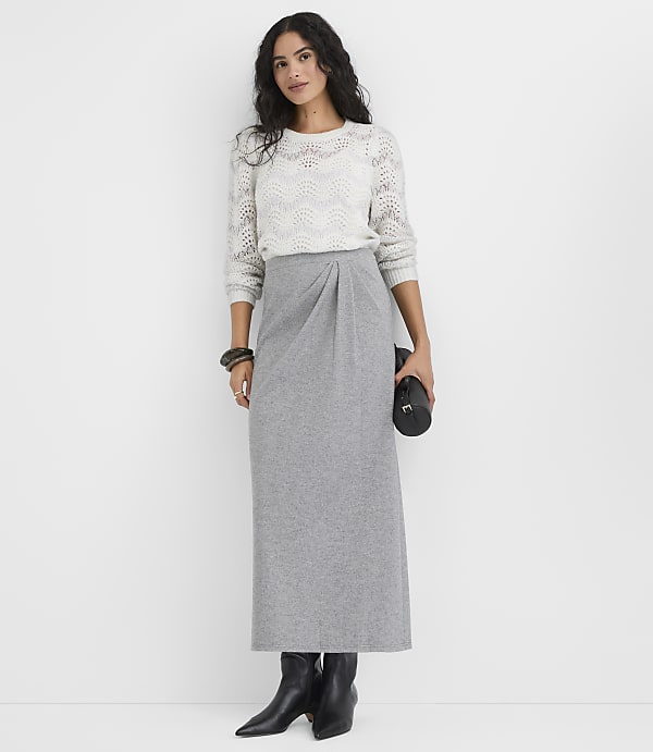 A woman wearing a white knit sweater and a long grey skirt stands against a plain white background.