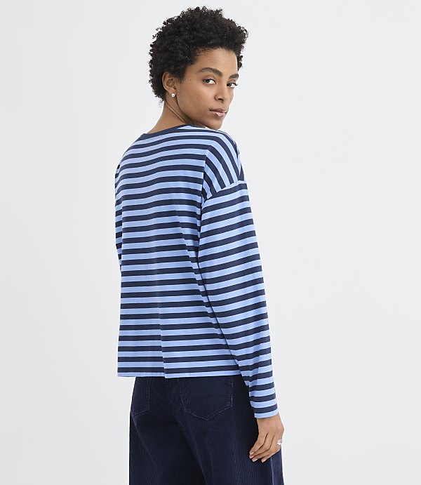 A woman wearing a blue and navy striped long-sleeve shirt and dark trousers stands against a plain white background.