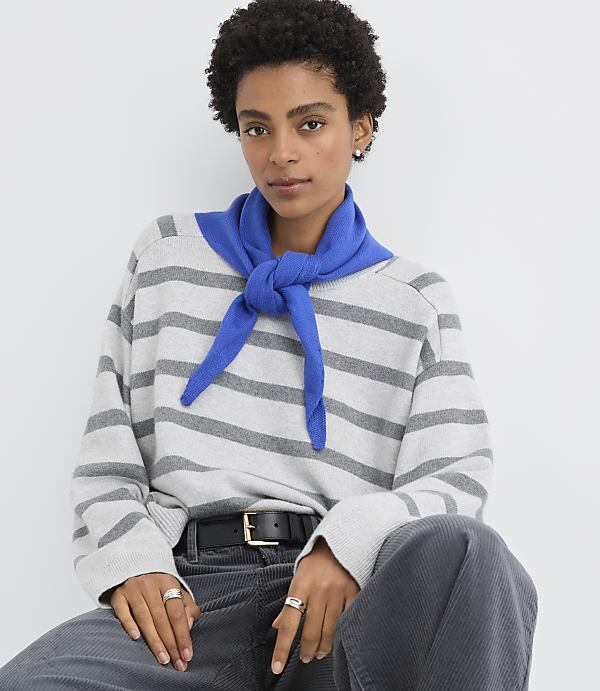 A woman wearing a grey striped sweater, a blue scarf, and dark trousers poses against a plain white background.