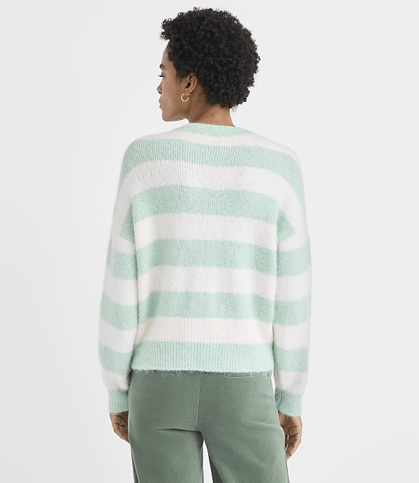 A woman with curly hair wearing a mint green and white striped sweater and green pants stands against a plain white background.