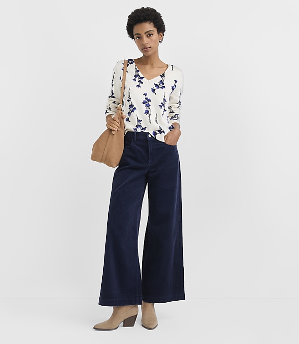 A woman wearing a floral white blouse and wide-leg navy trousers stands against a plain white background.