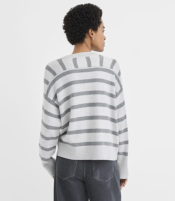 A woman wearing a grey and white striped sweater and dark grey pants stands against a plain white background.