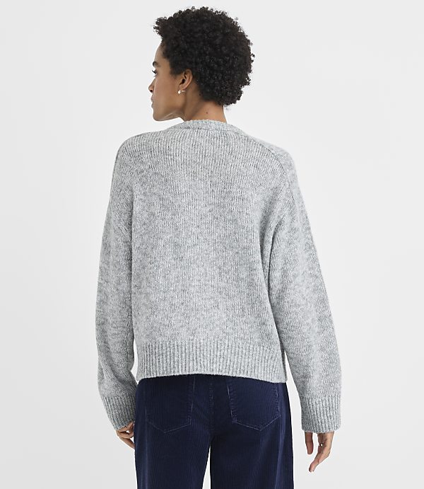 A woman wearing a grey knit sweater and navy blue corduroy pants stands against a plain white background.