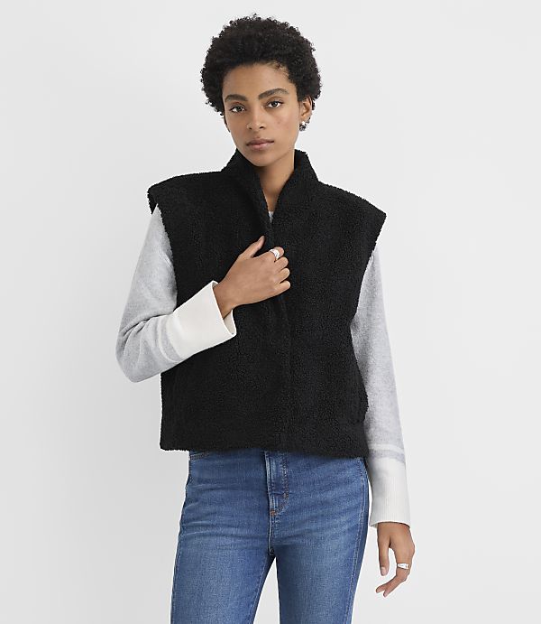 A woman wearing a black fleece vest over a grey long-sleeved shirt and blue jeans stands against a plain white background.