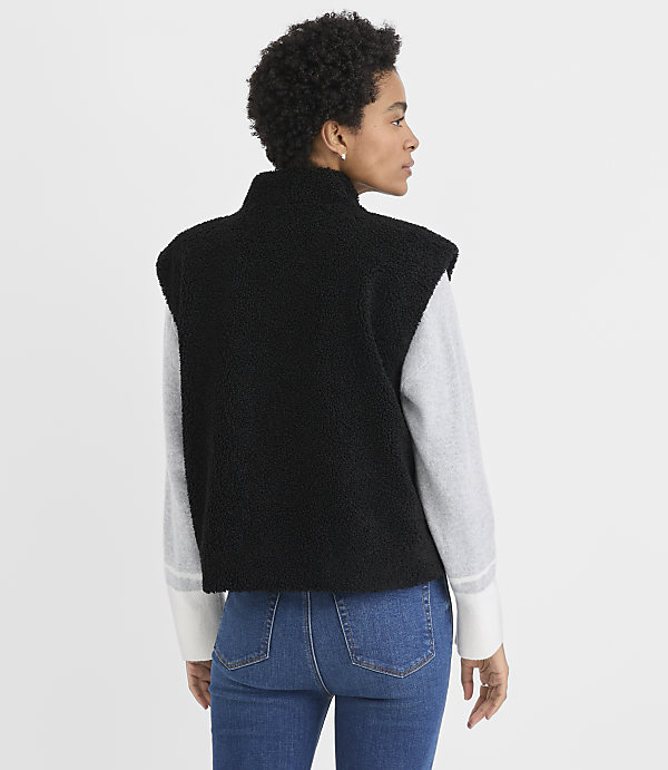 A woman wearing a black fleece vest over a grey long-sleeved shirt and blue jeans stands against a plain white background.