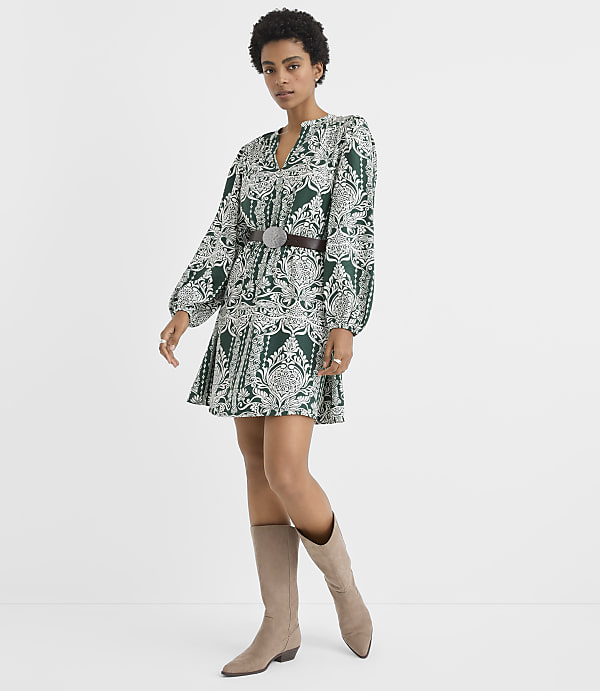 A woman wearing a green and white patterned mini dress with a belt and beige boots stands against a plain white background.