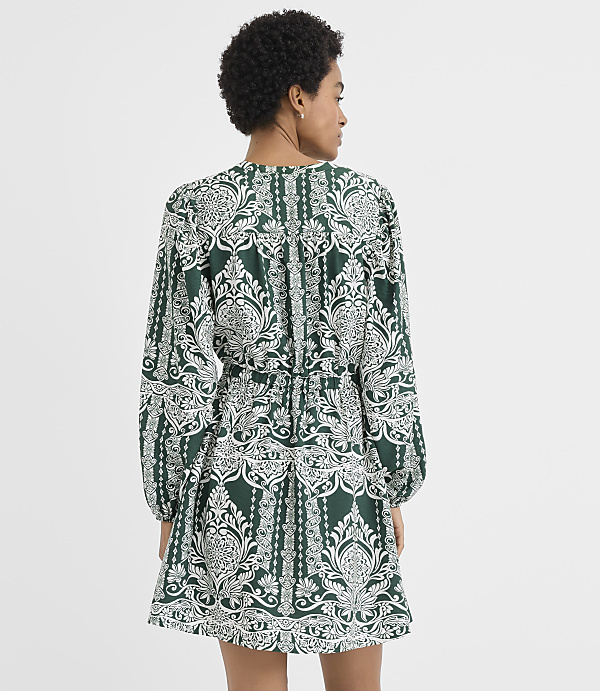 A woman wearing a green and white patterned dress stands with her back to the camera against a plain white background.