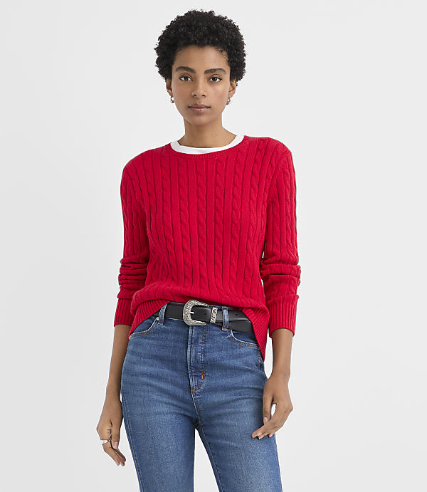 A woman wearing a red cable-knit sweater and blue jeans stands against a plain white background.