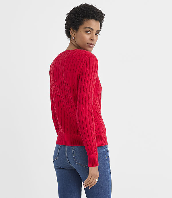 A woman wearing a red cable-knit sweater and blue jeans stands against a plain white background.