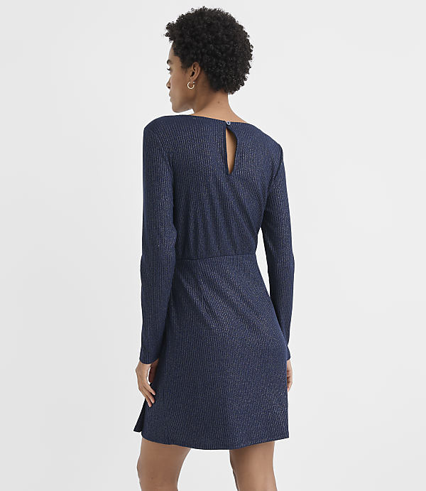 A woman with curly hair wearing a long-sleeved navy blue ribbed dress stands against a plain white background.