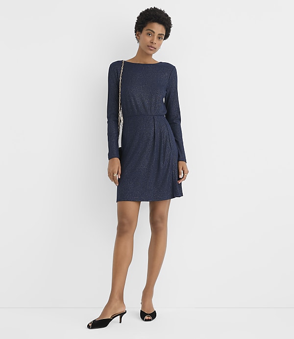 A woman wearing a navy blue long-sleeved dress and black heels stands against a plain white background.
