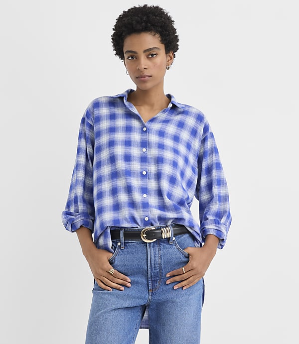 A woman wearing a blue plaid shirt and jeans stands against a plain white background.