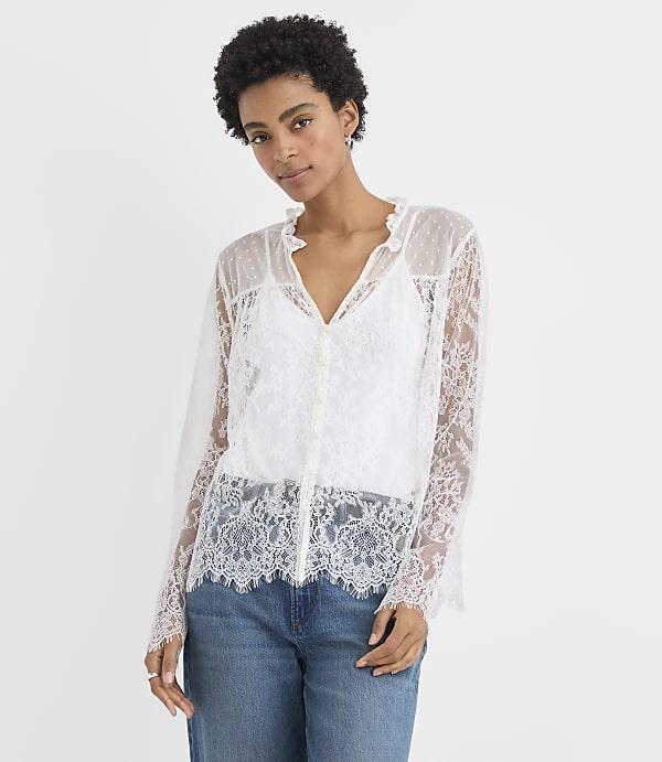 A woman wearing a white lace blouse and blue jeans stands against a plain white background.
