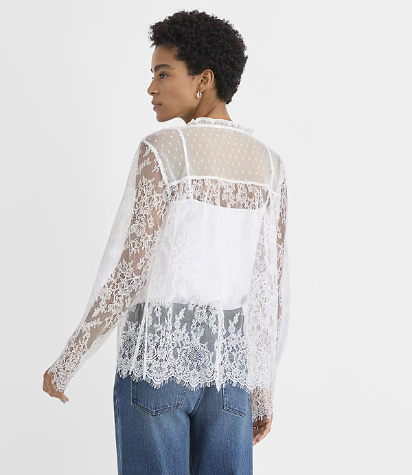 A woman wearing a white lace top and blue jeans stands against a plain white background.