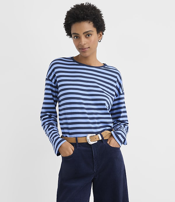 A woman wearing a blue and navy striped long-sleeve shirt and dark blue corduroy pants stands against a plain white background.