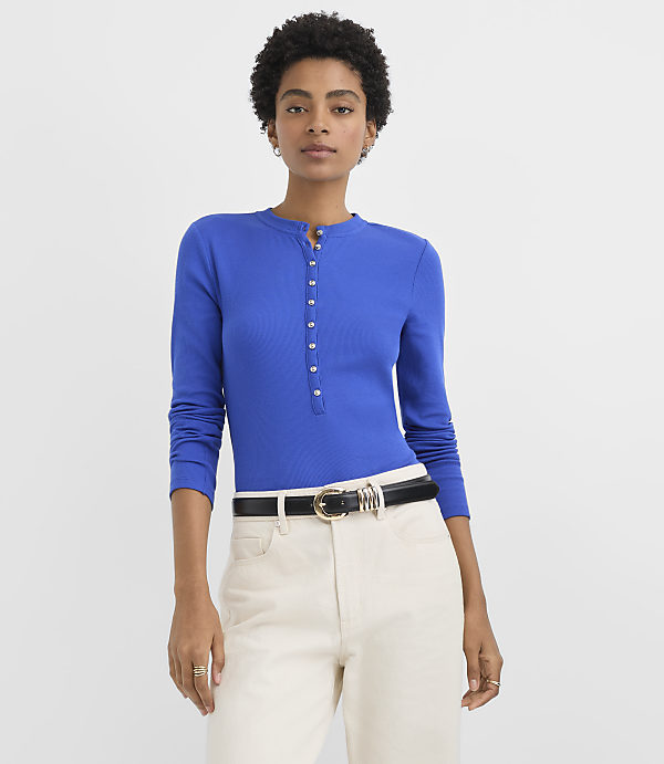 A woman wearing a royal blue button-placket top and cream-colored trousers stands against a plain white background.