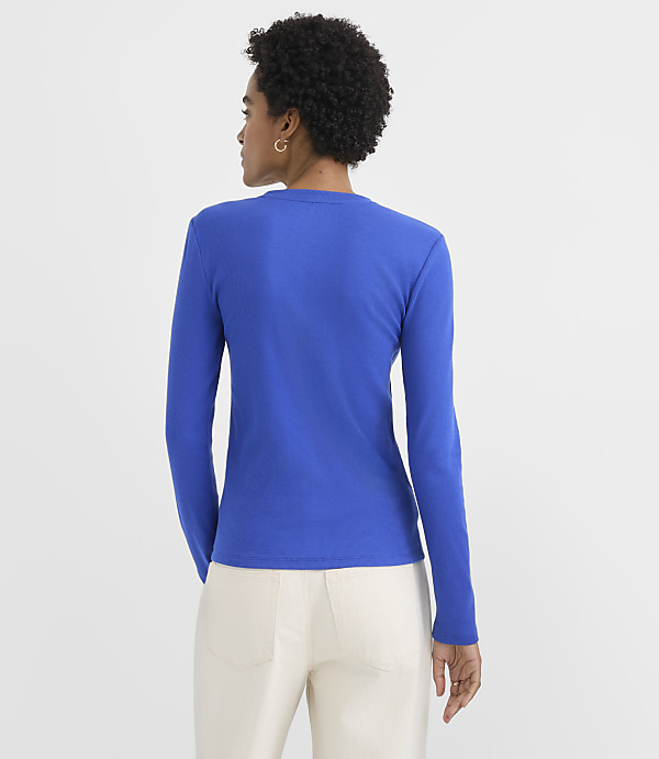 A woman wearing a bright blue long-sleeved shirt and white pants stands with her back to the camera against a plain white background.
