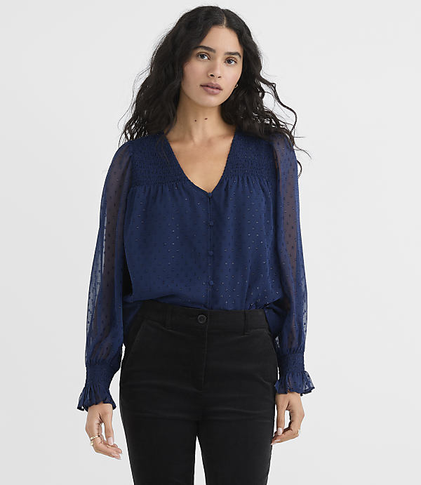 A woman wearing a navy blue sheer blouse and black trousers stands against a plain light gray background.