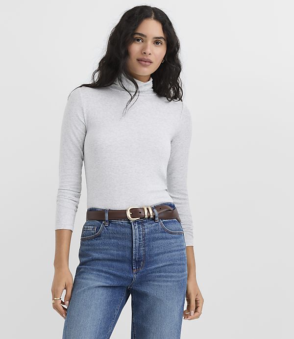 A woman wearing a grey turtleneck and blue jeans with a brown belt stands against a plain white background.