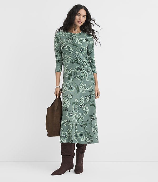A woman wearing a green floral midi dress and brown boots stands against a plain white background while holding a brown handbag.