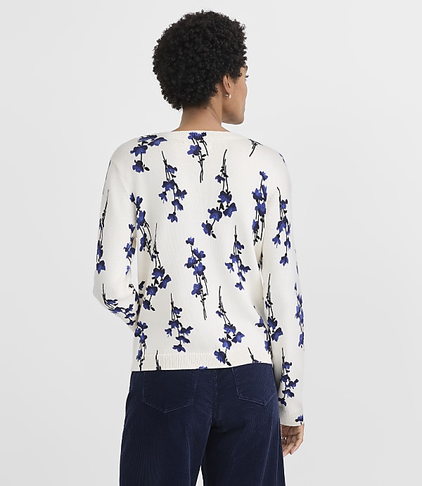 A woman with curly hair wearing a white floral sweater and dark blue pants stands against a plain light gray background.