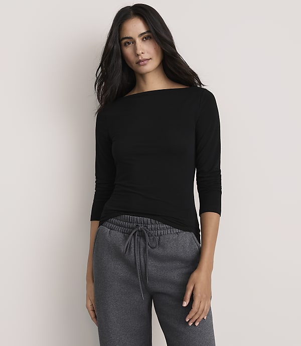 A woman wearing a black long-sleeved shirt and grey sweatpants stands against a plain, light-colored background.