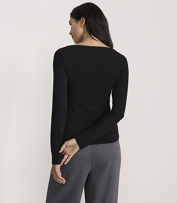 A woman wearing a black long-sleeved shirt and grey pants stands with her back to the camera against a plain white background.