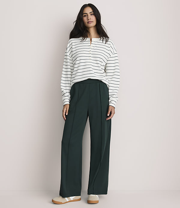 A woman wearing a striped long-sleeve shirt and wide-leg dark green trousers stands against a plain, light-colored background.