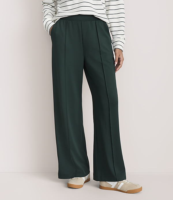 A person wearing a striped shirt, dark green wide-leg trousers, and beige sneakers stands against a plain, light-colored background.