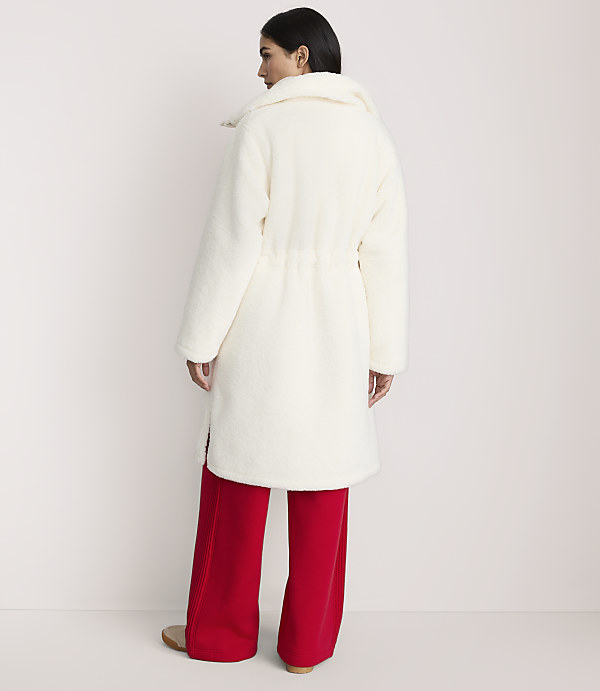 A woman wearing a long white faux-fur coat and wide-leg red trousers stands against a plain white background.