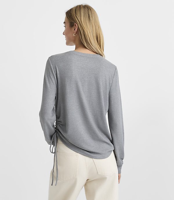 A woman wearing a grey long-sleeved shirt and white pants stands against a plain white background.
