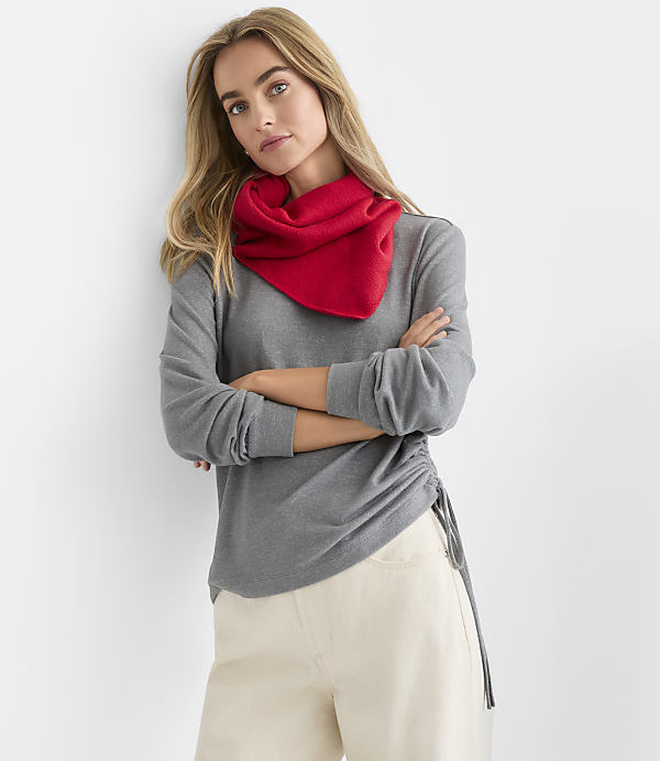 A blonde woman wearing a red scarf, grey long-sleeved shirt, and white pants stands with her arms crossed against a plain white background.