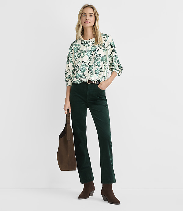 A woman wearing a patterned blouse and dark green corduroy pants stands against a plain white background.